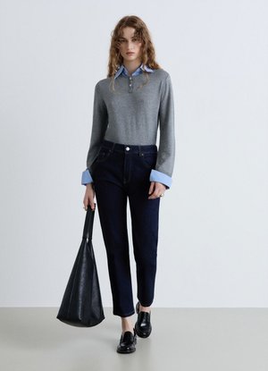 Donna con capelli ricci indossa un maglione grigio sopra una camicia blu, jeans scuri, scarpe nere e tiene una grande borsa di pelle nera, in piedi su un pavimento grigio.