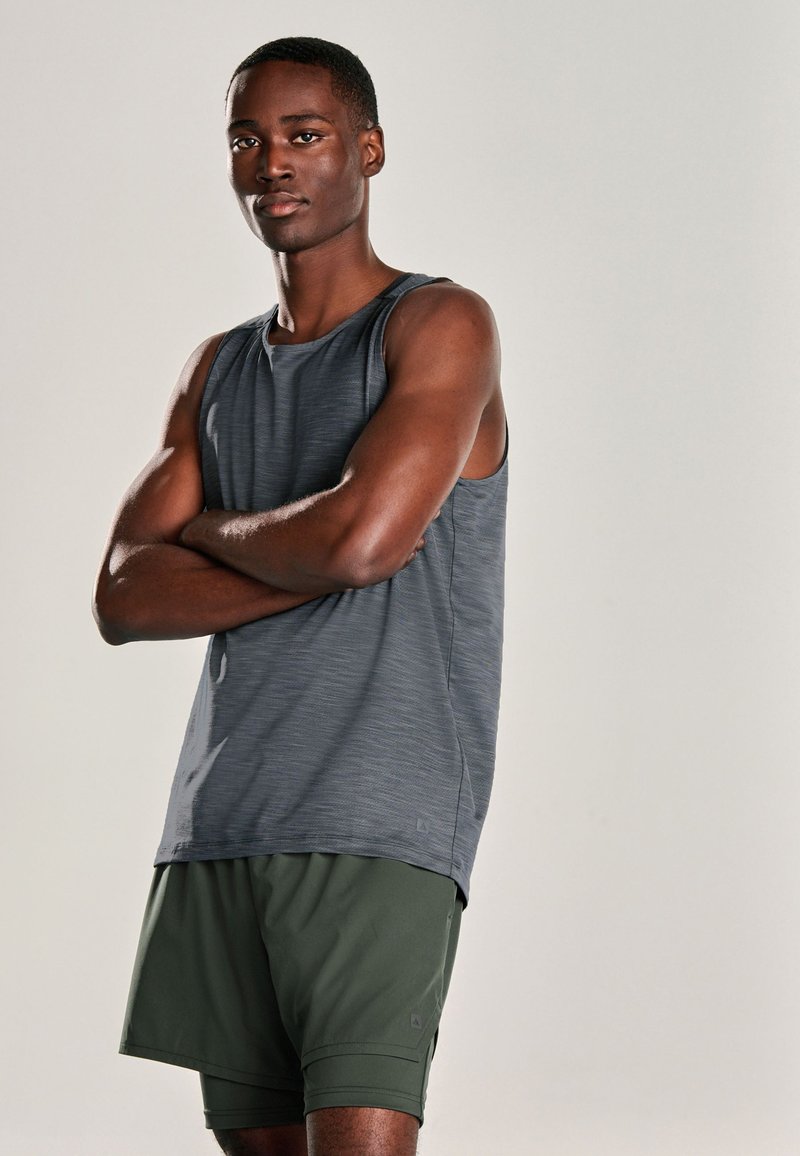 Homme bras croisés portant un haut de sport sans manches gris foncé et un short vert foncé, debout devant un fond uni.