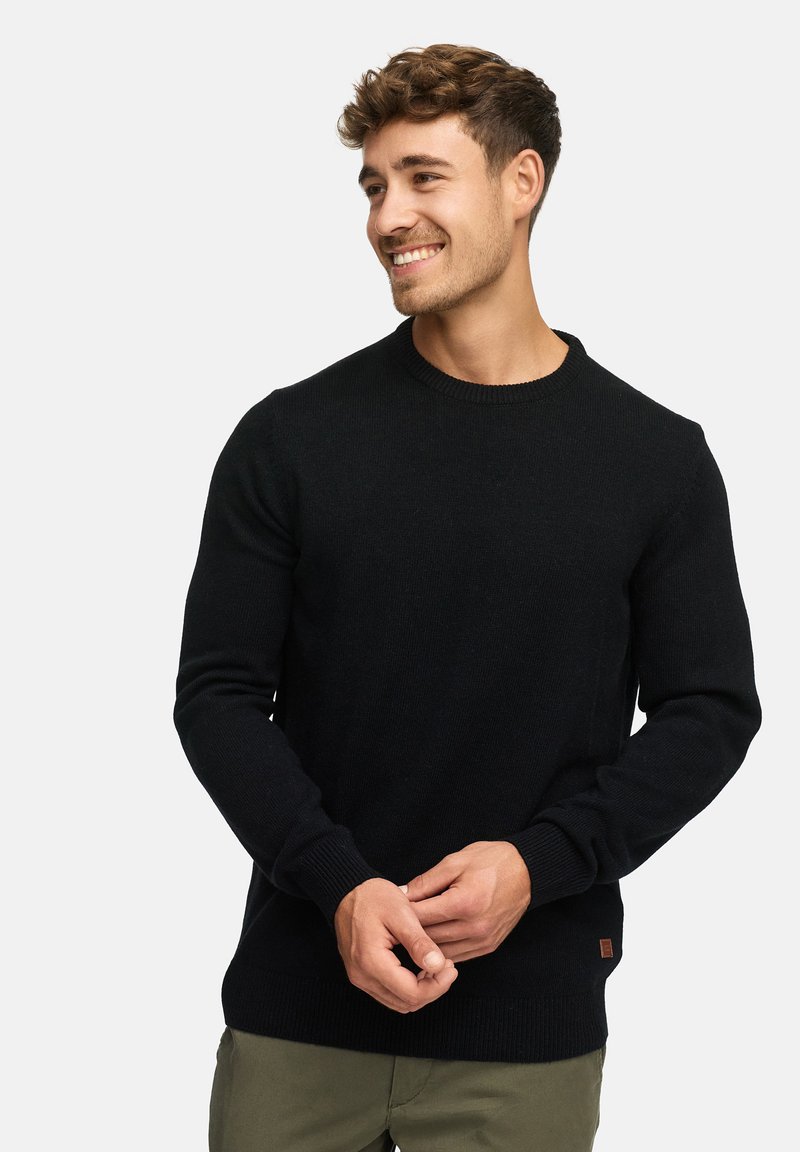 Pull en maille noir avec un col rond et des poignets côtelés. Doté d'un petit patch en cuir marron à l'ourlet. Texture lisse.