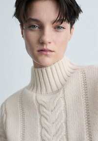 Cremfarbener Strickpullover mit hohem geripptem Kragen, Kabelmustern und strukturierten Streifen am Körper. Weicher, dicker Stoff.