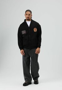 Zwarte varsity-cardigan met een sjaalkraag, knopen aan de voorkant, zijzakken, geborduurde applicaties en een losse pasvorm, gecombineerd met donkere, ruime spijkerbroeken.