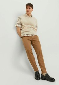 Gilet in maglia beige sopra una camicia a righe con bottoni, abbinato a pantaloni slim-fit marroni e mocassini neri. Texture morbide e tonalità neutre.
