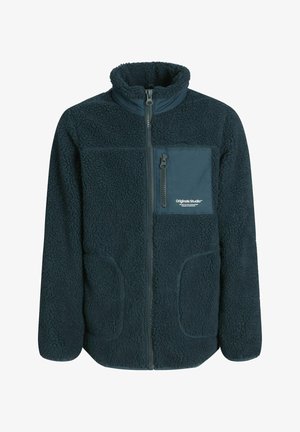 Giacca in pile verde scuro con collo alto, tasca sul petto con zip, indossata sopra una maglietta nera, abbinata a jeans azzurri e sneakers nere.