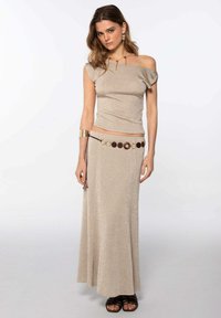 Top beige con brillo, con los hombros al descubierto, combinado con una falda larga y fluida de color beige. Acentuado con un cinturón circular marrón y joyería metálica.