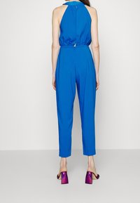 Blue halter jumpsuit with elastic waist and back button closure. Fitted silhouette with tapered legs. Sparkly purple block-heeled mules.