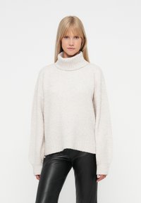 Maglione a collo alto beige chiaro con una finitura morbida e testurizzata, maniche ampie e orlo leggermente accorciato. Abbinato a pantaloni in pelle nera.