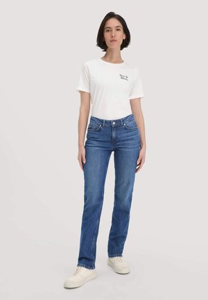 Vrouw in een wit t-shirt met de tekst "tijd om te bloeien", blauwe jeans en witte sneakers, staand tegen een eenvoudige lichte achtergrond.