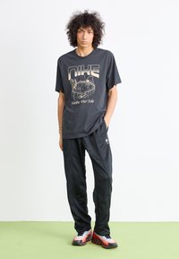 Ung person med krøllet hår iført sort Nike T-shirt, sorte træningsbukser og rød-hvide sneakers, som står på grøn gulv mod en hvid væg.