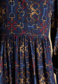 Tumesinine kleit, millel on tekstureeritud kangas kuldsete ja punaste abstraktsete mustritega, nööbilukk ja kogutud vöökoht.
