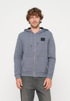 Junger Mann mit lockigem Haar, der einen blauen Kapuzenpullover mit Reißverschluss und einem schwarzen Aufnäher auf der Brust sowie graue Jeans vor einfachem Hintergrund trägt.