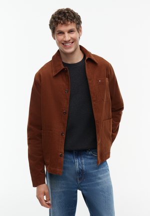 Ung mand smilende iført en brun kordfløjl jakke, sort sweater og blå jeans mod en ensfarvet hvid baggrund.