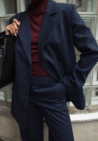 Ženska v mornarsko modrem kostimu in burgundski turtlenecku drži črno usnjeno torbico ter stoji ob steni iz steklenih blokov.