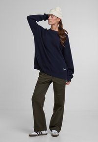 Pullover in maglia blu navy oversize con maniche lunghe, abbinato a pantaloni ampi verde oliva e sneakers bianche, completato da un berretto in maglia color crema.