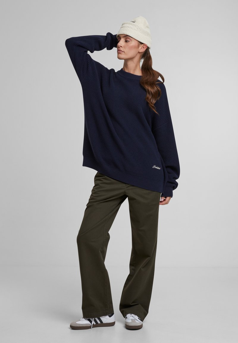 Pullover in maglia blu navy oversize con maniche lunghe, abbinato a pantaloni ampi verde oliva e sneakers bianche, completato da un berretto in maglia color crema.