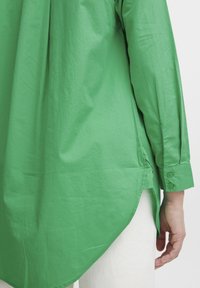 Green cotton shirt with long sleeves, a slightly curved hem, and buttoned cuffs. The fabric is smooth with a matte finish.
