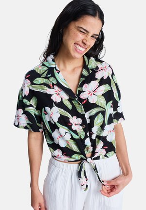 Frau zwinkert und lächelt, trägt ein schwarzes, kurzärmeliges Hemd mit Blumenmuster, das an der Taille gebunden ist, und lockere weiße Hosen vor weißem Hintergrund.