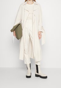 Vrouw in een crèmekleurige trenchcoat, met hoge taille broeken en plateau-laarzen, terwijl ze een grote olijfgroene clutch vasthoudt tegen een witte achtergrond.