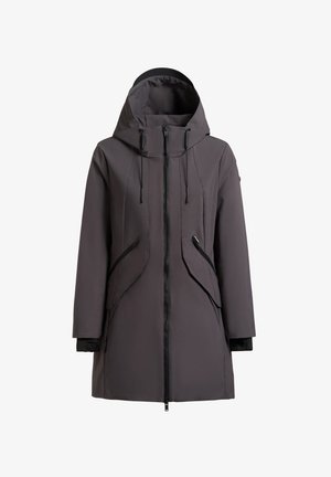 Žena stoji, nosi sivu kapuljaču, crne helanke i crne tenisice, na jednostavnoj svijetlosivoj pozadini.