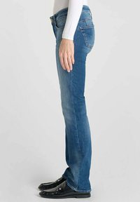 Jean en denim bleu avec une coupe droite, des coutures visibles, cinq poches et un léger délavage. Porté avec des chaussures en cuir verni noires.