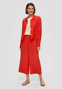 Veste en lin rouge avec des boutons dorés et une poche poitrine, associée à un pantalon large assorti. Un haut crème et des sandales dorées complètent la tenue.