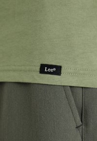 Tkanina oliwkowej koszulki z miękką fakturą i przeszytym dołem, z małą czarną metką z napisem "Lee®" na dolnym brzegu.