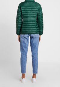 Grøn quiltet jakke med lodret syning, krave og lange ærmer, båret over lyseblå jeans og hvide sneakers, set bagfra.