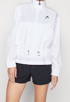 Person iført en hvid lynlåstrøje med elastiske ærmekanter og sorte shorts, stående mod en ensartet lys baggrund.