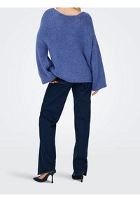 Femme portant un pull en maille bleu oversize, un jean droit bleu foncé et des chaussures à talons hauts bleu marine, debout dos à la caméra.