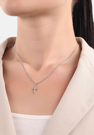 Frau trägt eine dünne silberne Kettenhalskette mit einem kleinen Kreuzanhänger, gekleidet in einem beigen Blazer und einem weißen Oberteil, mit zurückgebundenem Haar.