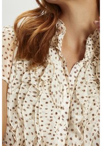 Blouse en crème translucent avec des pois marron, décolleté volanté et boutons sur le devant. Sa texture délicate et son tissu doux créent un design fluide.