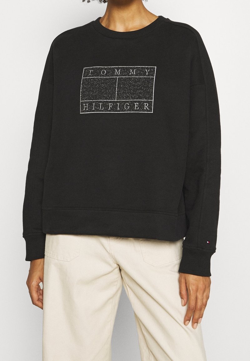 Personne portant un sweat-shirt noir Tommy Hilfiger avec logo blanc et un pantalon beige, debout devant un fond clair uni.
