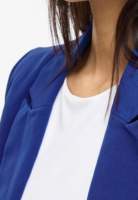 Femme portant un haut blanc et un blazer bleu, avec des cheveux châtain foncé tombant librement autour de son cou et de ses épaules.
