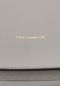 Grå läderplånbok med strukturerad yta, prydd med guldpräglad "COCCINELLE"-logotyp. Har synlig sömnad längs kanterna.