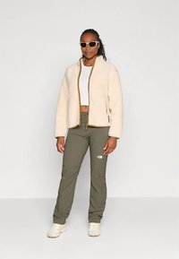 Veste en polaire crème avec fermeture éclair et bordure dorées, associée à un pantalon vert olive ; modèle portant un crop top blanc et des lunettes de soleil.