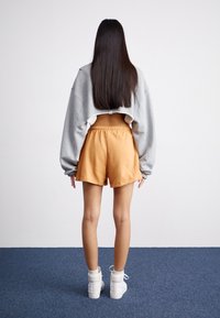 Szürke rövid fazonú sweatshirt, narancssárga sort, fehér sportcipő. Simaság, laza szabás, elasztikus derékpánt a sorton, kék szőnyegen áll.
