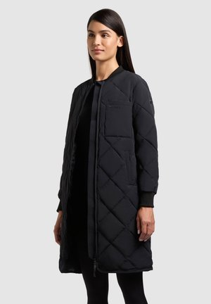 Femeie care poartă o haină lungă neagră matlasată, cu fermoar frontal, buzunar la piept, buzunare laterale și manșete striate, stând în fața unui fundal simplu.