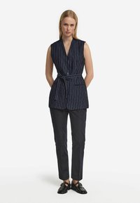 Marinblå ärmlös väst med vita randningar, en V-nacke, framknappar och midjebälte. Kombineras med smala mörka byxor och blanka svarta skor.