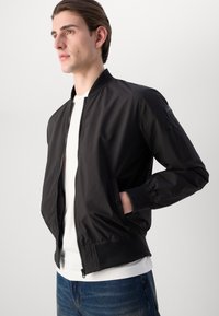 Schwarze Bomberjacke mit glatter, glänzender Oberfläche, gerippten Bündchen und einem frontalen Reißverschluss. Verfügt über Seitentaschen und eine zusätzliche mit Reißverschluss versehenen Ärmeltasche.
