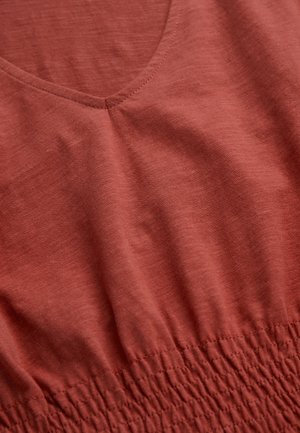 Roestkleurig kledingstuk met V-hals en geplooide elastische details in de taille, waarbij de stoftextuur en het stiksel zichtbaar zijn.