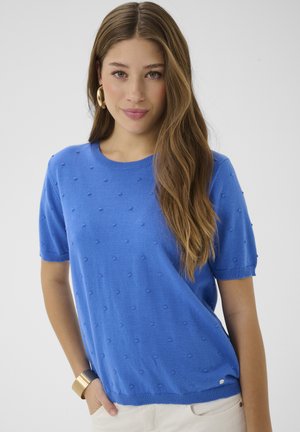 Ženska z dolgimi rjavimi lasmi, oblečena v modri, teksturiran pulover s kratkimi rokavi, bele hlače, zlatimi obročkastimi uhani in zlatim zapestnim trakom.