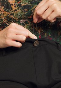 Pantalon noir avec une fermeture à bouton sur le devant, montrant des mains ajustant un cordon de serrage. Le tissu semble lisse avec un design de taille ajustée.