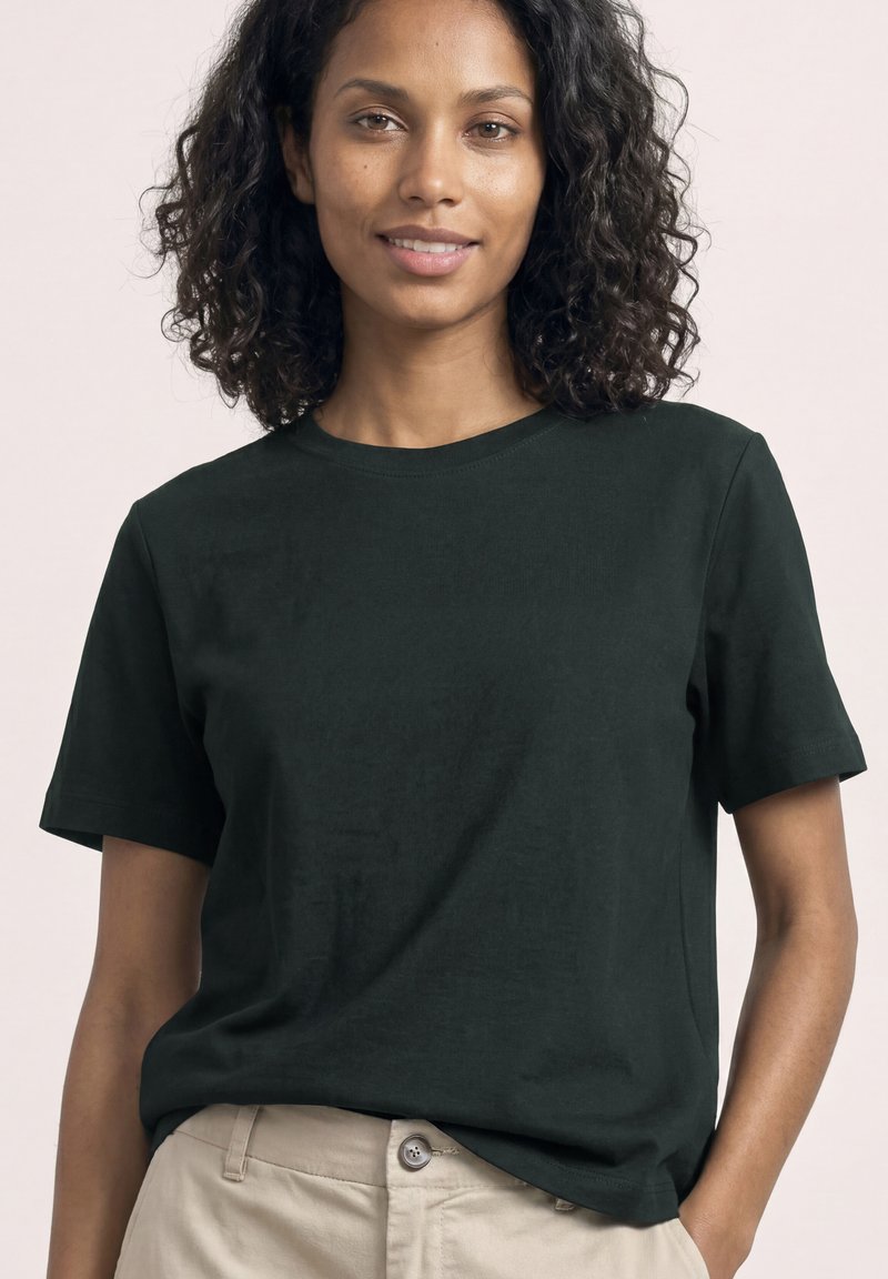 Femme aux cheveux bouclés et foncés porte un t-shirt noir uni à manches courtes et un pantalon beige, debout avec une main dans la poche, souriant doucement.