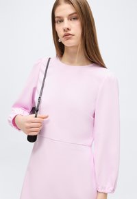 Jeune femme portant une robe à manches longues rose clair, tenant une bandoulière de sac en cuir noir sur son épaule sur un fond uni.