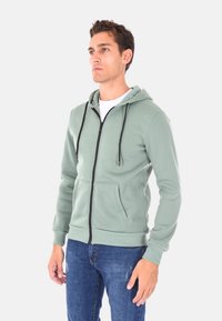Felpa verde con zip, dotata di una tasca frontale, stringhe nere e polsini a coste, abbinata a jeans blu. Tessuto liscio, design casual.