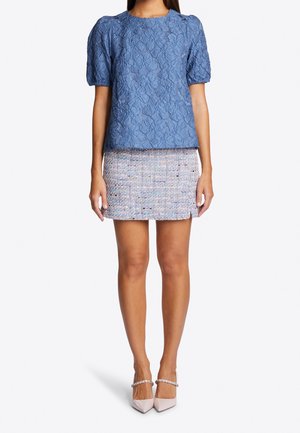 Mujer con blusa azul de manga corta y textura, minifalda de tweed multicolor con una pequeña abertura y zapatos de tacón puntiagudos rosa pálido con tiras en el tobillo.