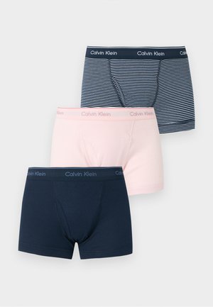 Bărbat slab cu pielea închisă la culoare, stând desculț pe un fundal simplu, purtând chiloți boxer Calvin Klein cu dungi albastre și albe.