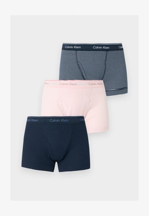 Vékony, sötét bőrű férfi áll mezítláb egyszínű háttér előtt, kék-fehér csíkos Calvin Klein boxeralsót viselve.