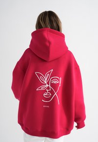 Felpa rossa in tessuto morbido, con un design in linea bianca di un volto e foglie sul retro, con una vestibilità ampia e rilassata.