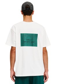 Weißes Baumwoll-T-Shirt mit einem grünen, geometrischen Grafikdesign und dem Text "HALO" sowie "OFF DUTY GEAR", sichtbar von hinten.