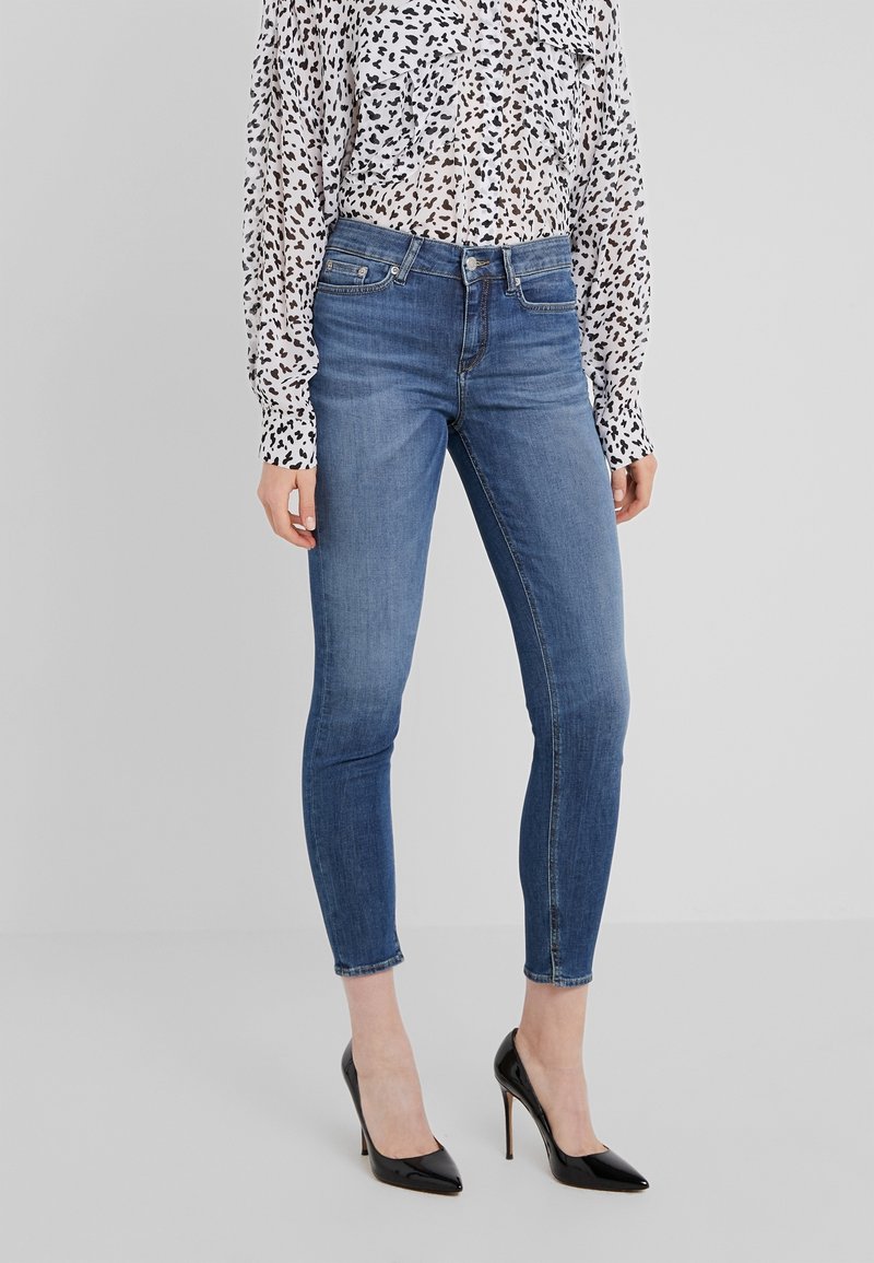 Jeans ajustados azules con cintura de altura media, con un ligero desvanecimiento y diseño de cinco bolsillos, combinados con una blusa blanca con estampado y tacones altos negros.